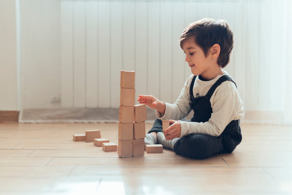 A child sits on the floor, focused on building a tower with wooden blocks