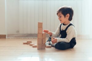 A child sits on the floor, focused on building a tower with wooden blocks