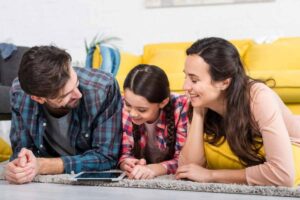 A family of three lies on a carpet, watching a tablet together