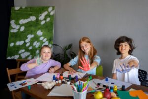 Three children smiling, showing painted hands, while surrounded by art supplies and a colourful painting in the background.
