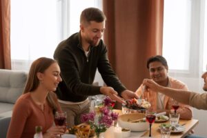 A man serves food to friends at a dinner table with wine and flowers.