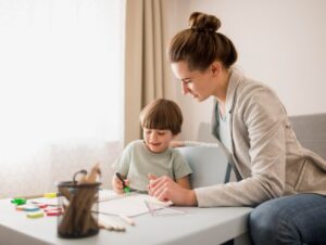 A young woman helps a small child with drawing at a white table, with art supplies scattered around.