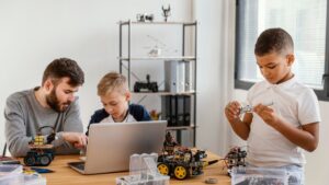 An adult and two children engaging with robots and a laptop on a desk.