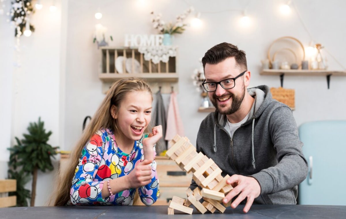 A man and a young girl laugh together as they play with wooden blocks at a cosy home setup.