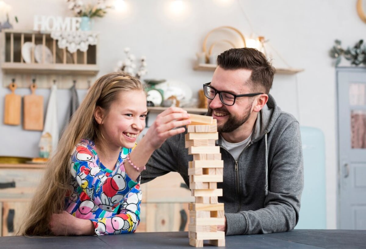 A man and a young girl smiling as they play with a wooden block tower in a cozy kitchen setting.