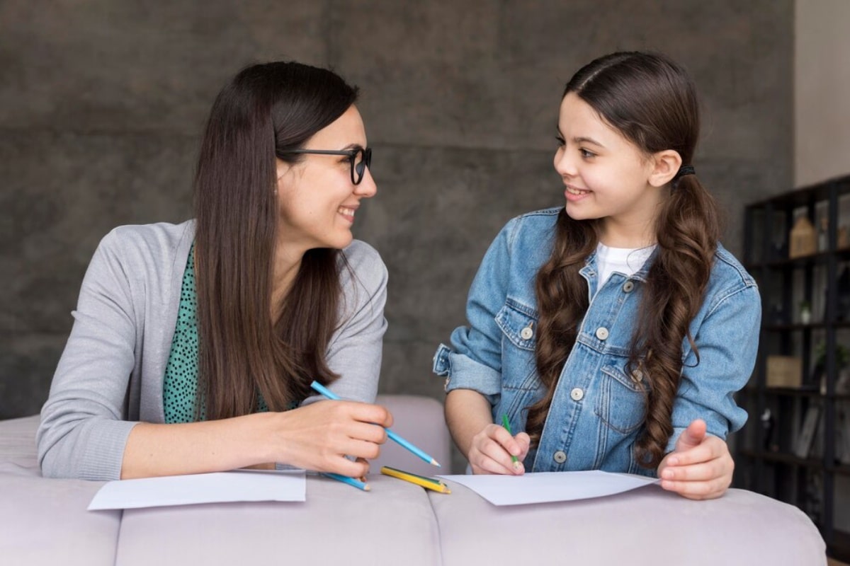 A woman in glasses and a girl in a denim jacket smiling and writing at a table.