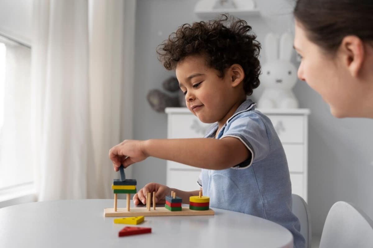 Young boy playing with colorful wooden blocks at a table, with a woman watching him.