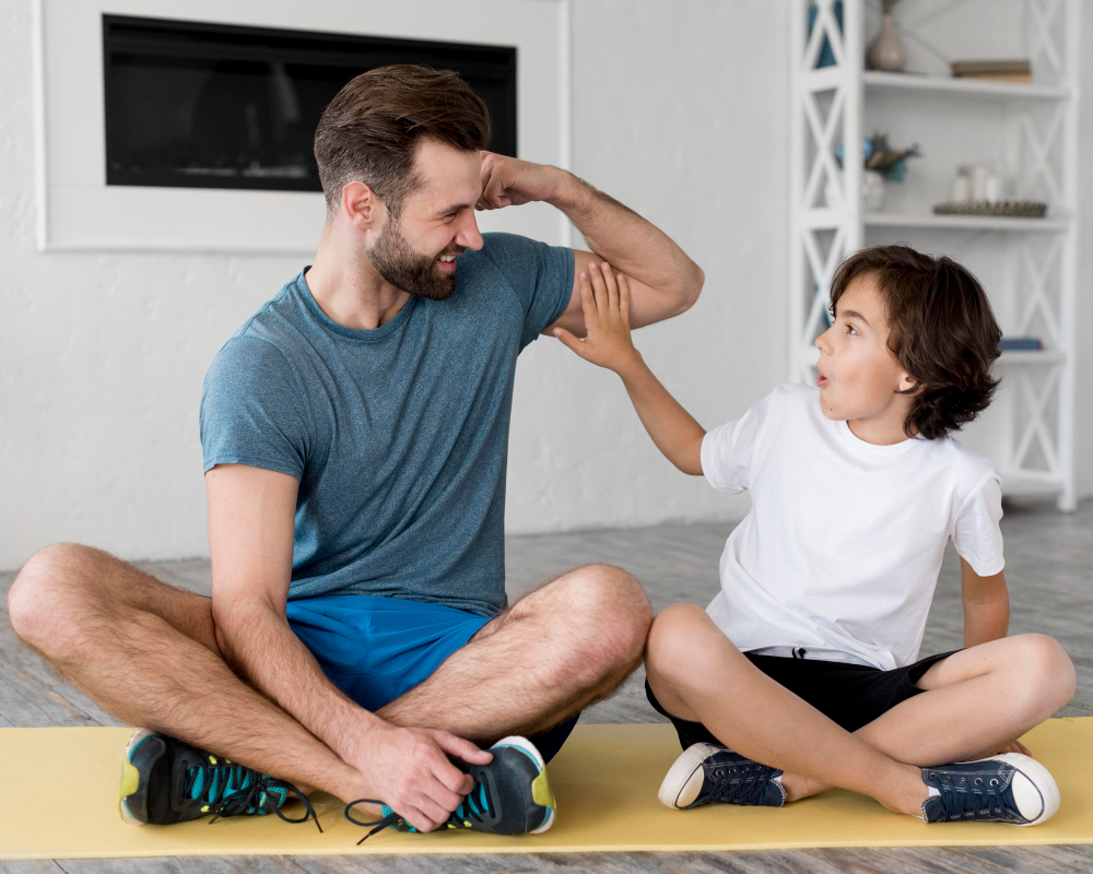 A man and a child are seated on a yoga mat, with the man flexing his arm while the child touches it playfully.