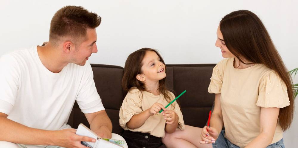 A family sits together on a couch, with a young girl playfully holding colorful pens, engaged in a warm conversation.